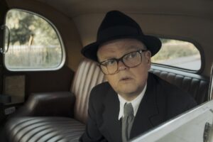 Toby Jones as Phillip Burton wears a suit and trilby in an old fashioned car. He peers through the window.